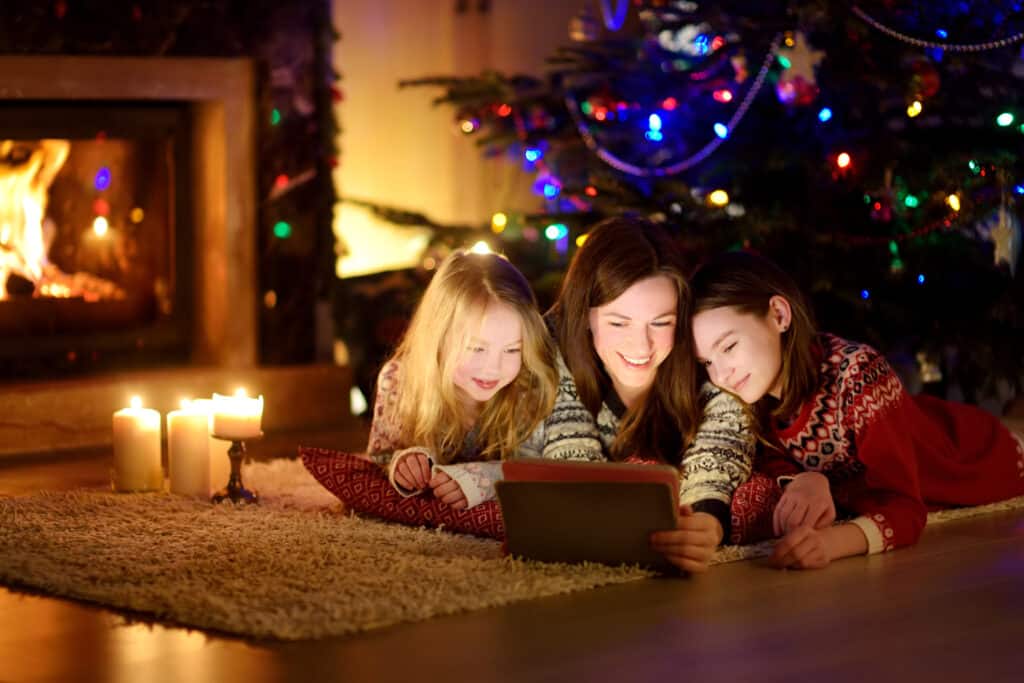 Mother and her two cute young daughters using a tablet pc at home by a fireplace in warm and cozy living room on Christmas eve. Winter evening at home with family and kids.